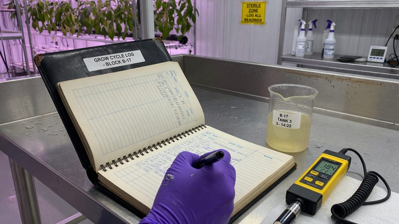 Gloved hand writing in a bound logbook on a stainless bench beside an EC meter